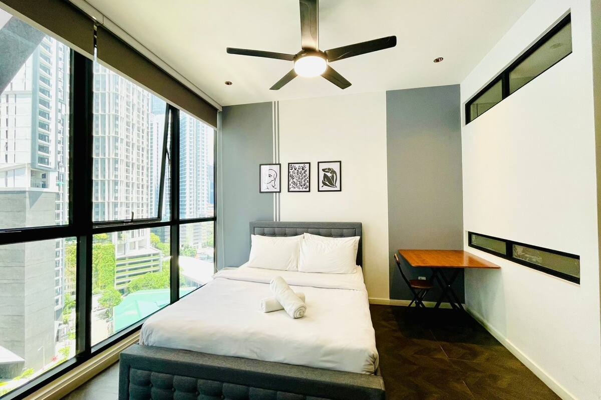 A modern hotel room with a large bed, a ceiling fan, a small table, and a view of the city through large windows.