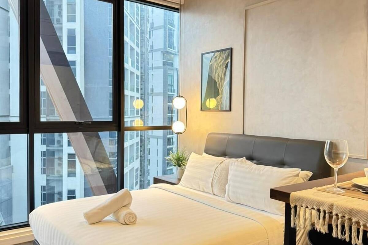 A modern hotel room with a large bed, a ceiling fan, a desk with a chair, and a large window offering a city view.
