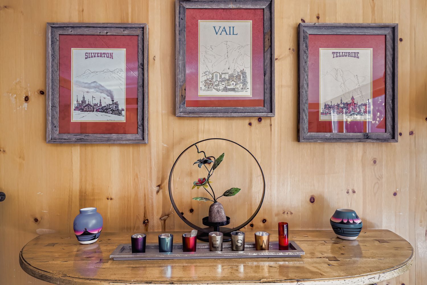 A wooden wall with three framed pictures labeled 'SILVERTON', 'VAIL', and 'TELLURIDE'. Below, a round metal frame holds a decorative plant.