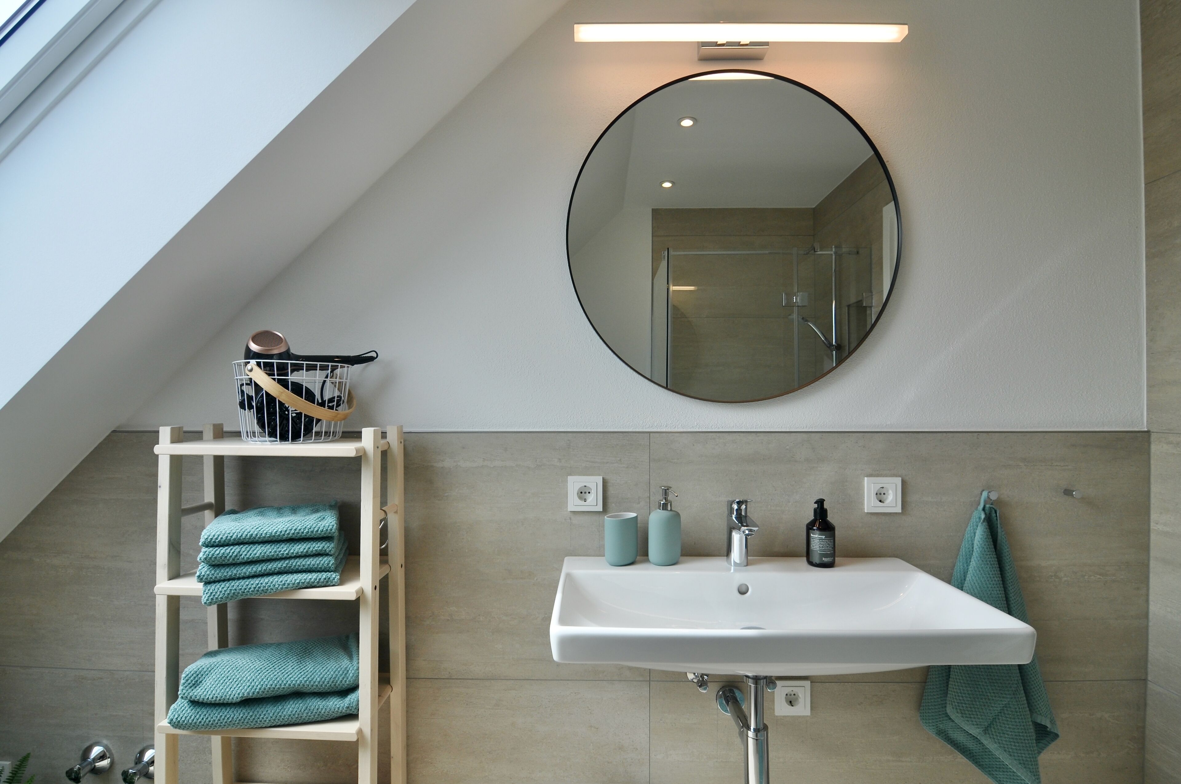 Un baño moderno con tragaluz, un espejo redondo, un lavabo blanco y un estante de madera con toallas.