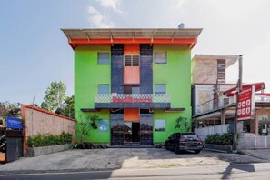 Un edificio de dos pisos con fachada verde, entrada negra y un estacionamiento al frente.