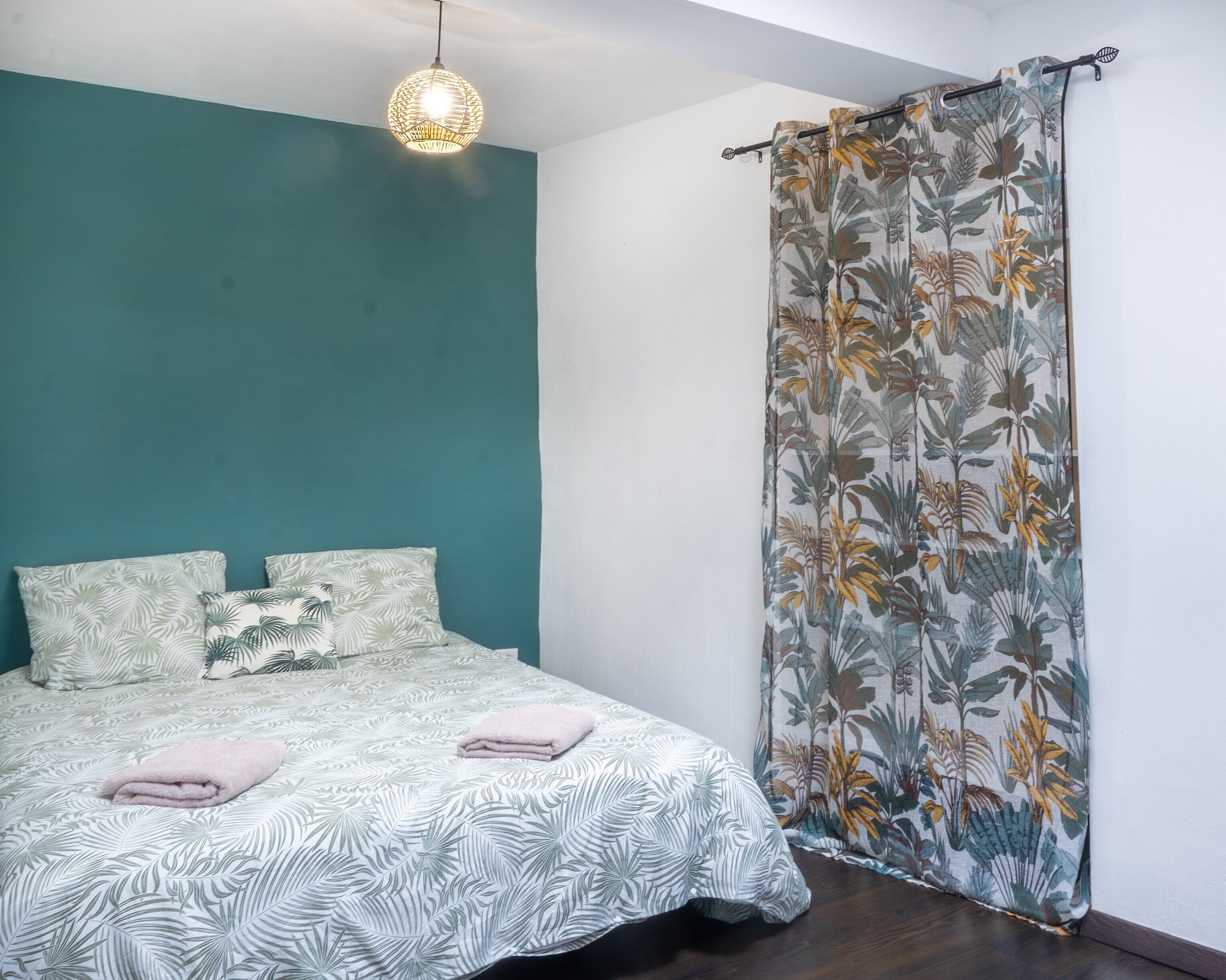 Een slaapkamer met een blauwgroene wand, een bed met beddengoed met een patroon, een hanglamp en een bloemen gordijn.