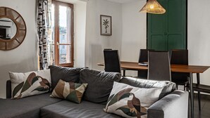 A modern living room with a sofa, a dining table, chairs, a hanging light fixture, a green door, and a window with patterned curtains. - Apartment 'La Glycine' with Wi-Fi and Air Conditioning (Montolieu)