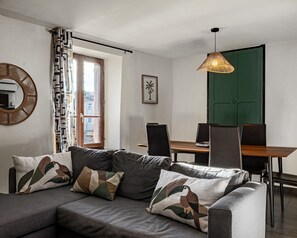 Uma sala de estar moderna com um sofá, uma mesa de jantar, cadeiras, uma luminária pendente, uma porta verde e uma janela com cortinas estampadas.