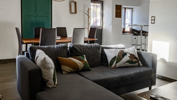 A living room with a grey sofa, a dining table with chairs, a hanging light fixture, and a green door. - Apartment 'La Glycine' with Wi-Fi and Air Conditioning (Montolieu)