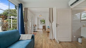 A room with a blue sofa, wooden flooring, a dining area with a table and chairs, and a view of greenery outside. - Villa Mandarine - Two Bedroom Villa, Sleeps 4 (Le Diamant)