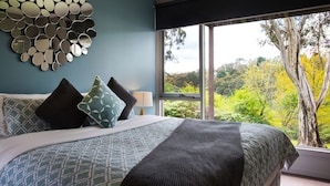 Une chambre à coucher avec un lit, des oreillers, un miroir mural et une fenêtre donnant sur des arbres.