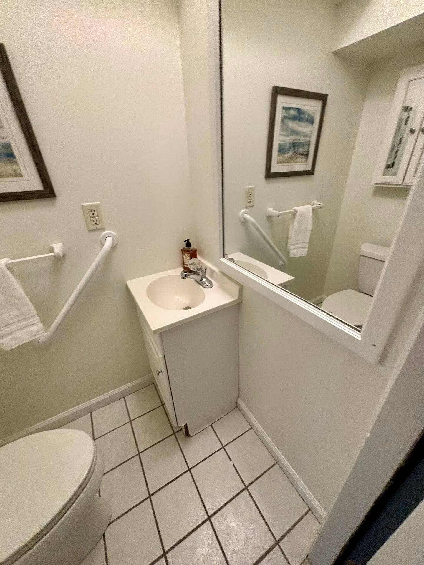 A small, white bathroom with a sink, mirror, and toilet. There is a handrail on the left side and a towel rack above the sink.