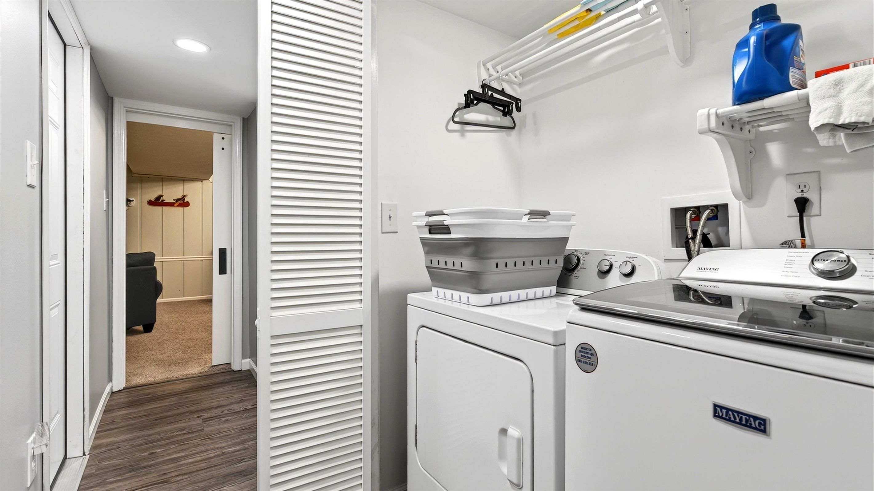 A laundry room with a Maytag washer and dryer, a drying rack, and a shelf with cleaning supplies.