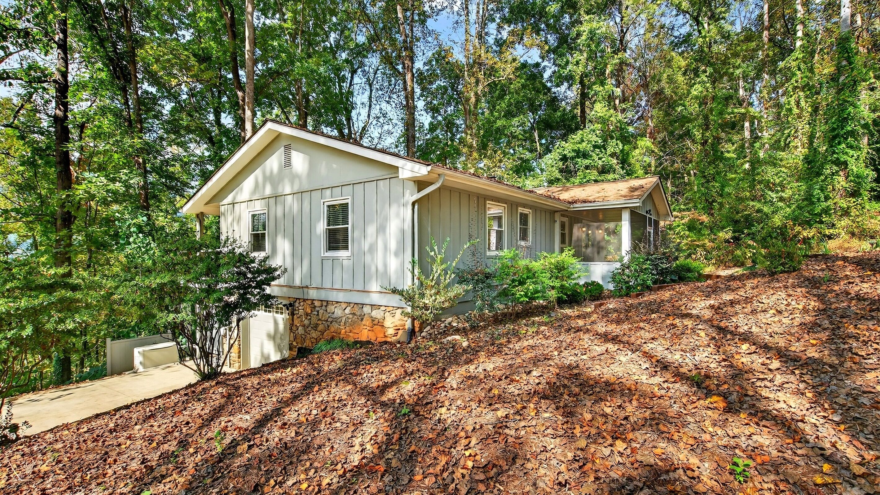 A house with a stone foundation, a front porch, and a garage, surrounded by dense forest and autumn leaves.