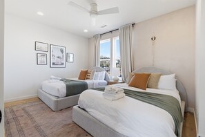 Ein Hotelzimmer mit zwei Betten, einem Deckenventilator, einem Fenster mit Vorhängen und gerahmten Bildern an der Wand.