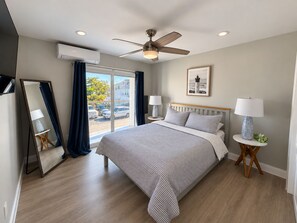 A hotel room with a bed, ceiling fan, air conditioning unit, two bedside lamps, a framed picture on the wall, and a view through the window.