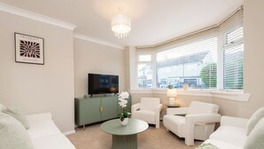 Un salón con un mueble verde, televisor, muebles blancos y una ventana con persianas.