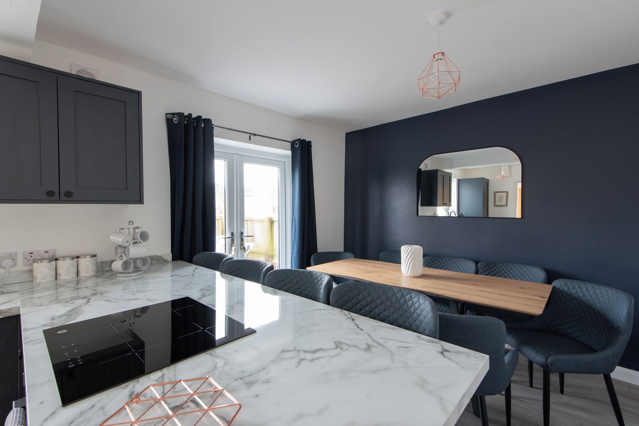 A modern kitchen with a marble countertop, dark blue upholstered chairs, and a wooden dining table.