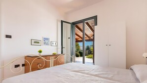 Ein Schlafzimmer mit Bett, Schminktisch, einem kleinen Tisch mit Pflanze und einer Tür, die auf einen Balkon führt.
