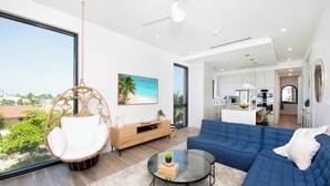 A modern living room with a large window, a blue sofa, a glass coffee table, a wicker chair, and a wooden cabinet. - SeaDreams 6: Oceanfront Condos w/Pool, Hot Tub, & Rooftop Patio (West Bay)