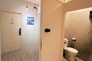 A bathroom with a toilet, shower, and a door leading to a hallway with a light fixture and a wall-mounted picture. - DaDa97 Hostel (Bangkok)