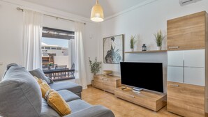Ein modernes Wohnzimmer mit Sofa, Fernseher, Holzschrank und einem durch eine Glastür einsehbaren Essbereich.
