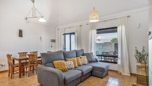 Ein Wohnzimmer mit einem grauen Ecksofa, einem Esstisch mit Stühlen und einem großen Fenster mit Vorhängen.