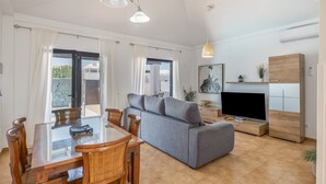 Ein modernes Wohnzimmer mit einem Glastisch, Holzsesseln, einem grauen Sofa, einem Fernseher und einer Holzkommode.