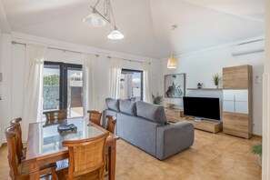 Un salon moderne avec une table à manger en verre, des chaises en bois, un canapé gris, un téléviseur et un meuble en bois.