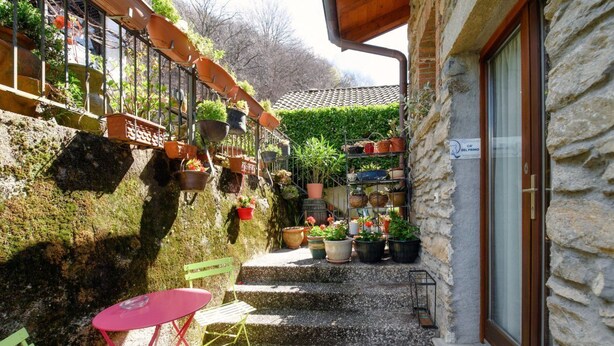 A stone building with a small outdoor seating area, steps leading to a door, and various potted plants. - Two-room apartment in the valleys of Capriasca (Ca' del Primo) (Odogno)