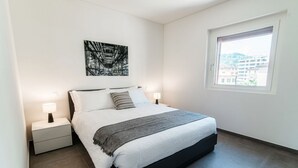Ett modernt sovrum med en säng, sängbord, ett fönster och utsikt över staden.