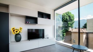 Ett modernt vardagsrum med en platt-TV, en vas med solrosor och ett matbord placerat vid ett stort fönster med utsikt över stadsbilden.