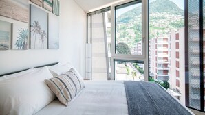 Un dormitorio con un ventanal grande, una cama con ropa de cama blanca y vistas a un paisaje urbano y colinas.