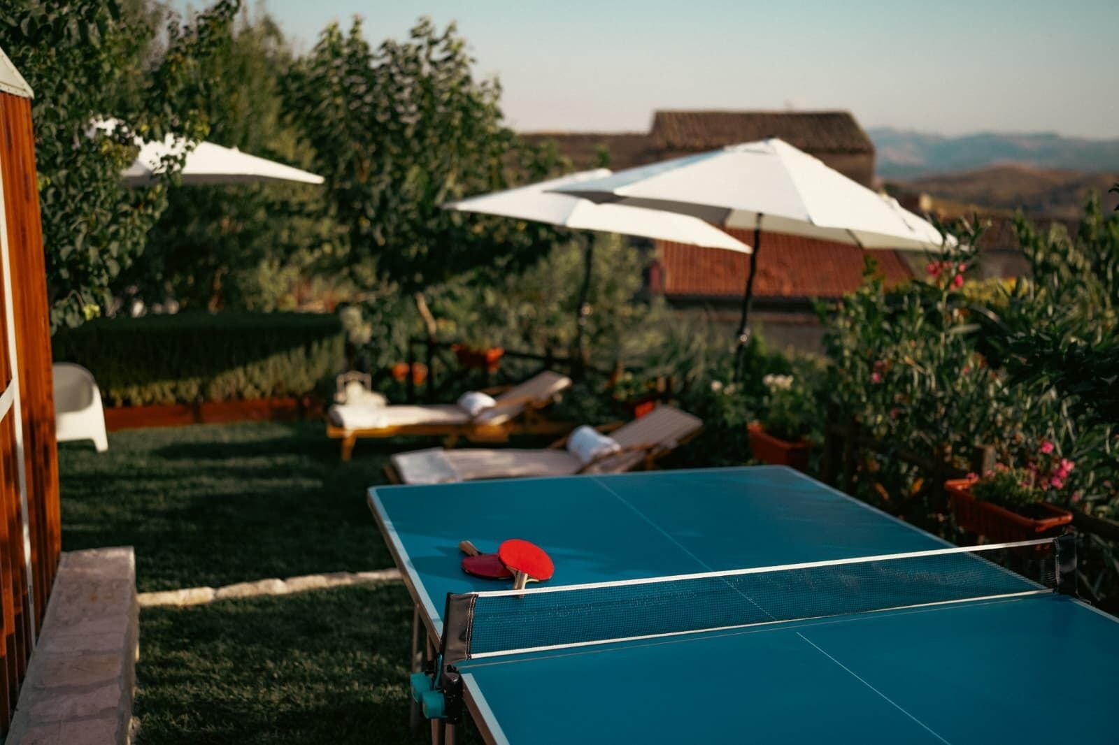 Ein Tischtennis-Tisch im Freien mit zwei weißen Sonnenschirmen, umgeben von Liegestühlen und Grünpflanzen.