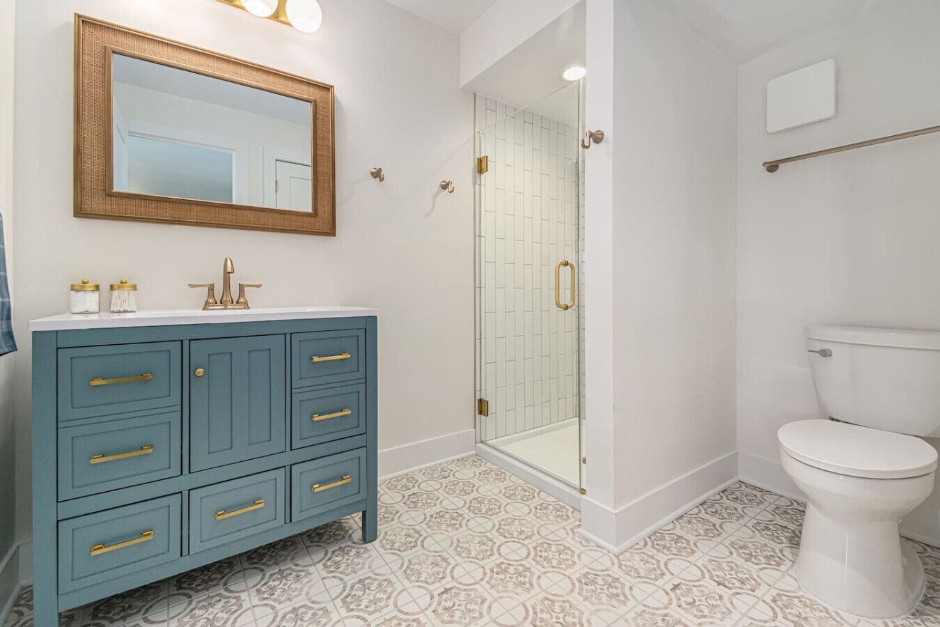 Un baño con inodoro blanco, tocador azul con tiradores dorados, cabina de ducha de vidrio y un espejo grande con marco de madera.