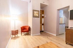 A room with a wooden floor, a red chair, a small wooden side table, a framed picture on the wall, and a closet with hanging clothes. - Stylish Aparthotel | 1 Room+Lounge by Mariahilfer (Vienna)