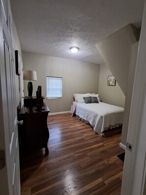 Ein Schlafzimmer mit Bett, Schreibtisch, Lampe, Fenster und Deckenleuchte.