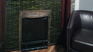 Une cheminée ornée de carreaux verts, un miroir décoratif au-dessus, et un fauteuil en cuir sur la droite.