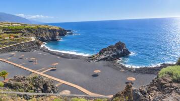 Un paisaje costero con playa rocosa, mar azul cristalino y montañas al fondo.