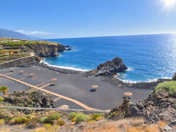 A coastal landscape with a rocky beach, clear blue sea, and a mountainous backdrop.