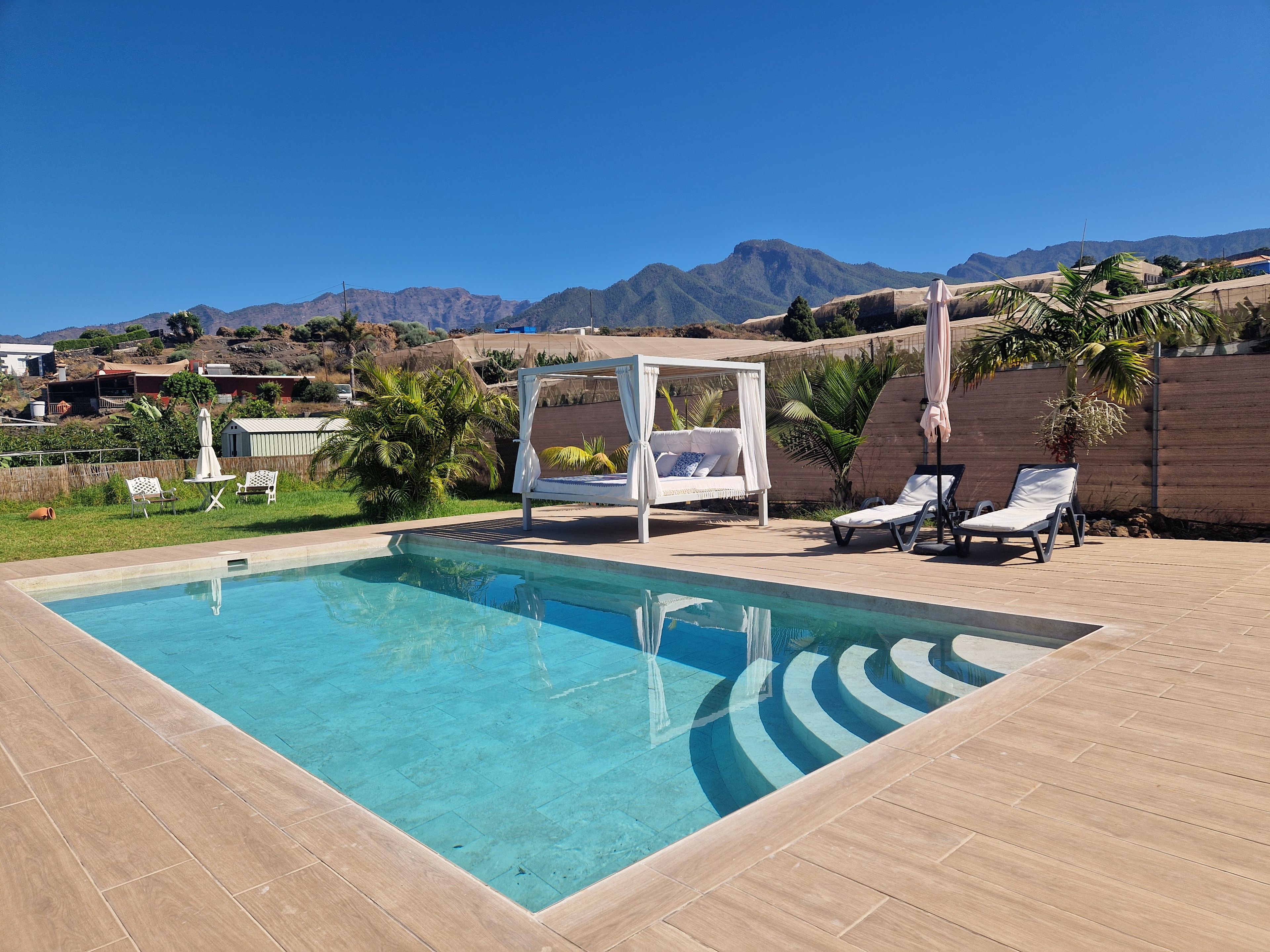 Una piscina con sillones de descanso y una sombrilla, rodeada por una terraza de madera y palmeras, con montañas al fondo.