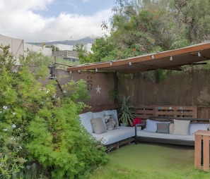 A covered outdoor seating area with a wooden bench and cushions, surrounded by lush greenery and a view of a mountainous landscape.