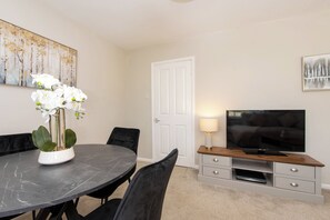 A dining area with a round table, chairs, a potted plant, a TV on a wooden stand, and artwork on the walls. - Toast Stays - Broomfield House (Durham)