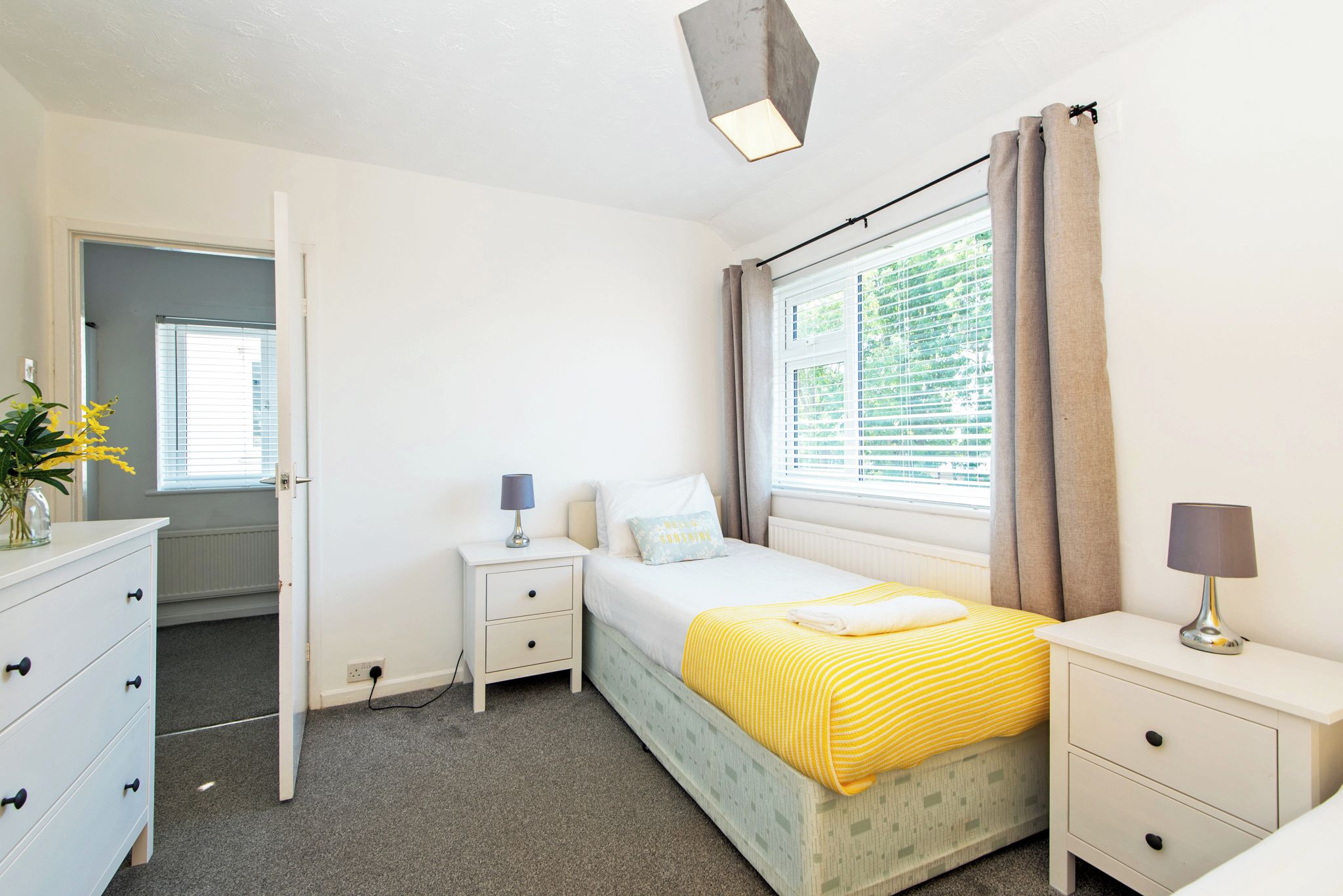 A bedroom with a bed, bedside tables, a dresser, a window with curtains, and a door leading to another room.