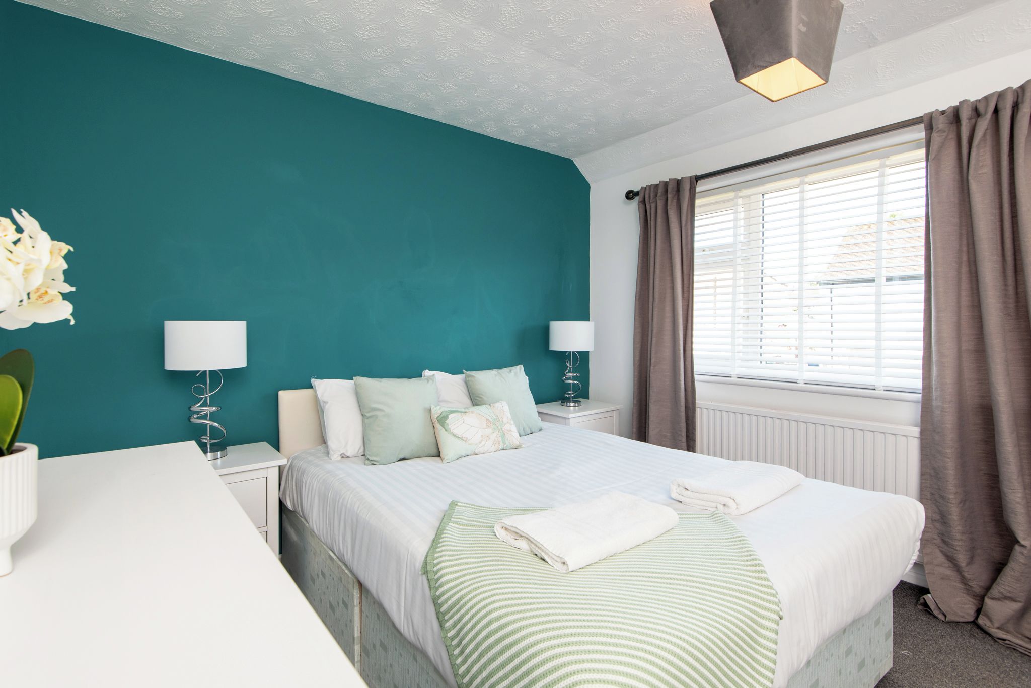 Een slaapkamer met turquoise wanden, een bed met witte lakens, twee bedlampjes, een raam met gordijnen en een bloempot met plant.