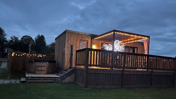 Una terraza de madera con una escalera que conduce a un área cubierta, rodeada por una valla de madera y luces colgantes.