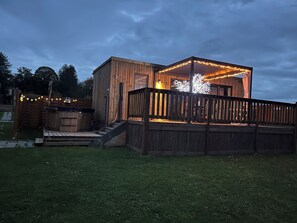 Une terrasse en bois avec un escalier menant à une zone couverte, le tout entouré d’une clôture en bois et illuminé de guirlandes lumineuses.