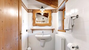 Ein Badezimmer mit Holztür, weißen Fliesen, einem Waschbecken mit Spiegel darüber, einem Handtuchhalter und einem Heizkörper.