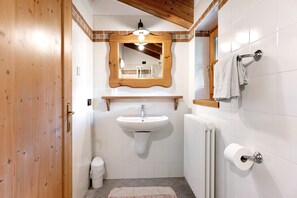 Un baño con puerta de madera, baldosas blancas, un lavamanos con espejo, un perchero y un radiador.