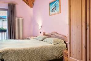 Un dormitorio con cama, mesita de noche, armario de madera, radiador, ventana con cortinas y un cuadro en la pared.