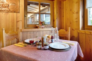 L’intérieur d’un chalet en bois présente une table à manger dressée pour deux personnes, avec une nappe à carreaux, des assiettes, des couverts et des pots de confitures.