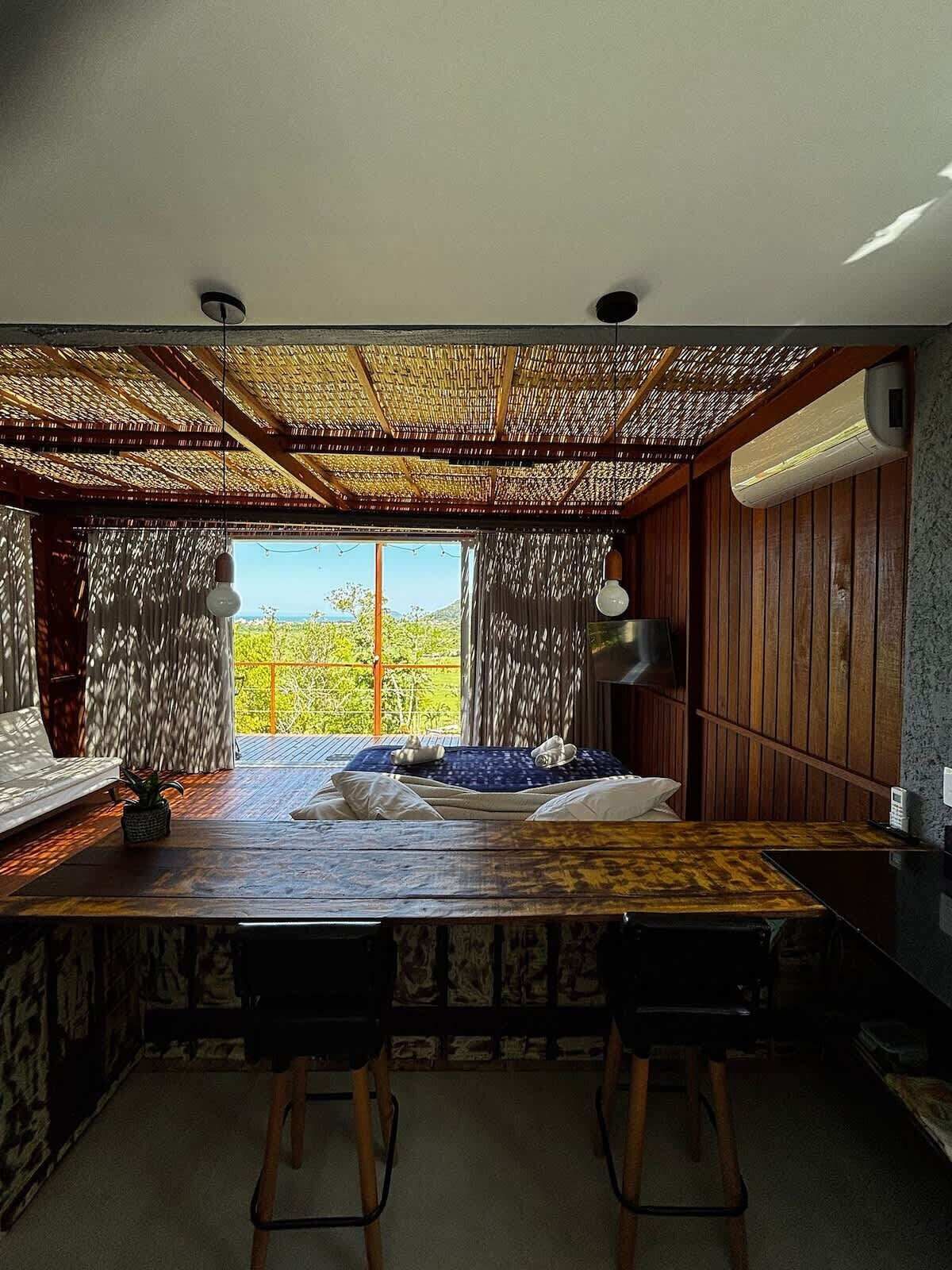 A room with a wooden ceiling, a large window, a bed, a table, and two chairs.