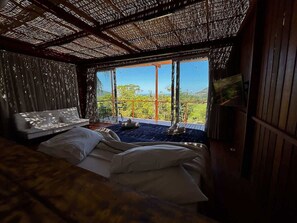 A room with a bed, a sofa, and a large window offering a view of greenery and hills.