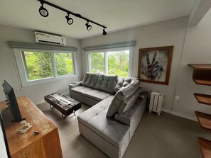 A modern living room with a sectional sofa, a small table, and a TV. There are large windows and a painting on the wall.